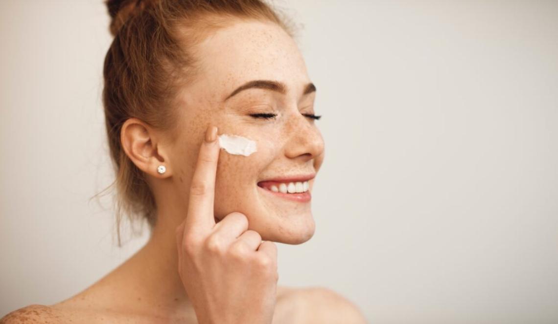 Close up of a young female with red hair and freckles applying white cream on her face laughing with closed eyes isolated on white background