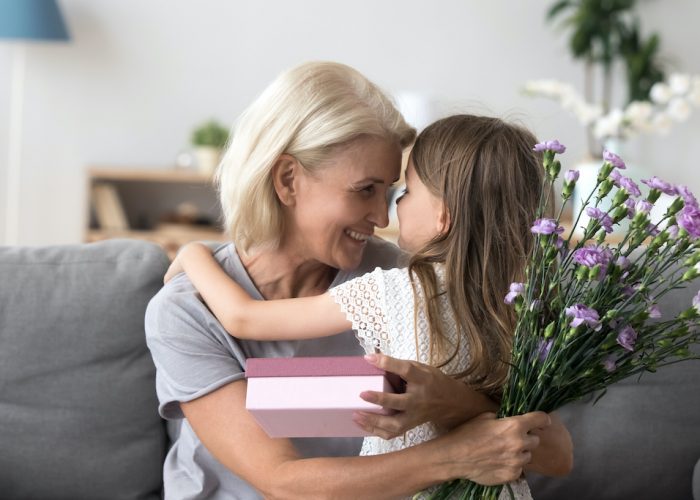 Happy grandmother and granddaughter hug celebrating birthday together, cute little girl congratulate granny presenting gift box and flowers bouquet, grandma thanking grandchild for surprise