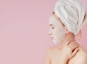 Beautiful young woman is applying a cosmetic tissue mask on a face on a pink background. Healthcare and beauty treatment and technology concept.