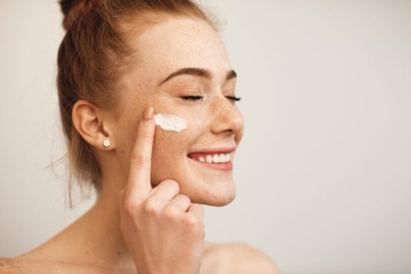 Close up of a young female with red hair and freckles applying white cream on her face laughing with closed eyes isolated on white background