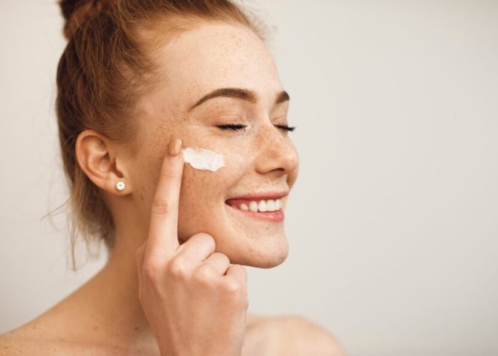 Close up of a young female with red hair and freckles applying white cream on her face laughing with closed eyes isolated on white background