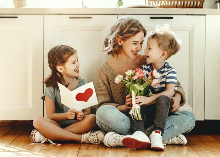 Young mother with a bouquet of tulips laughs, hugging her son, and  сheerful girl with a card congratulates mom during holiday celebration in kitchen at home